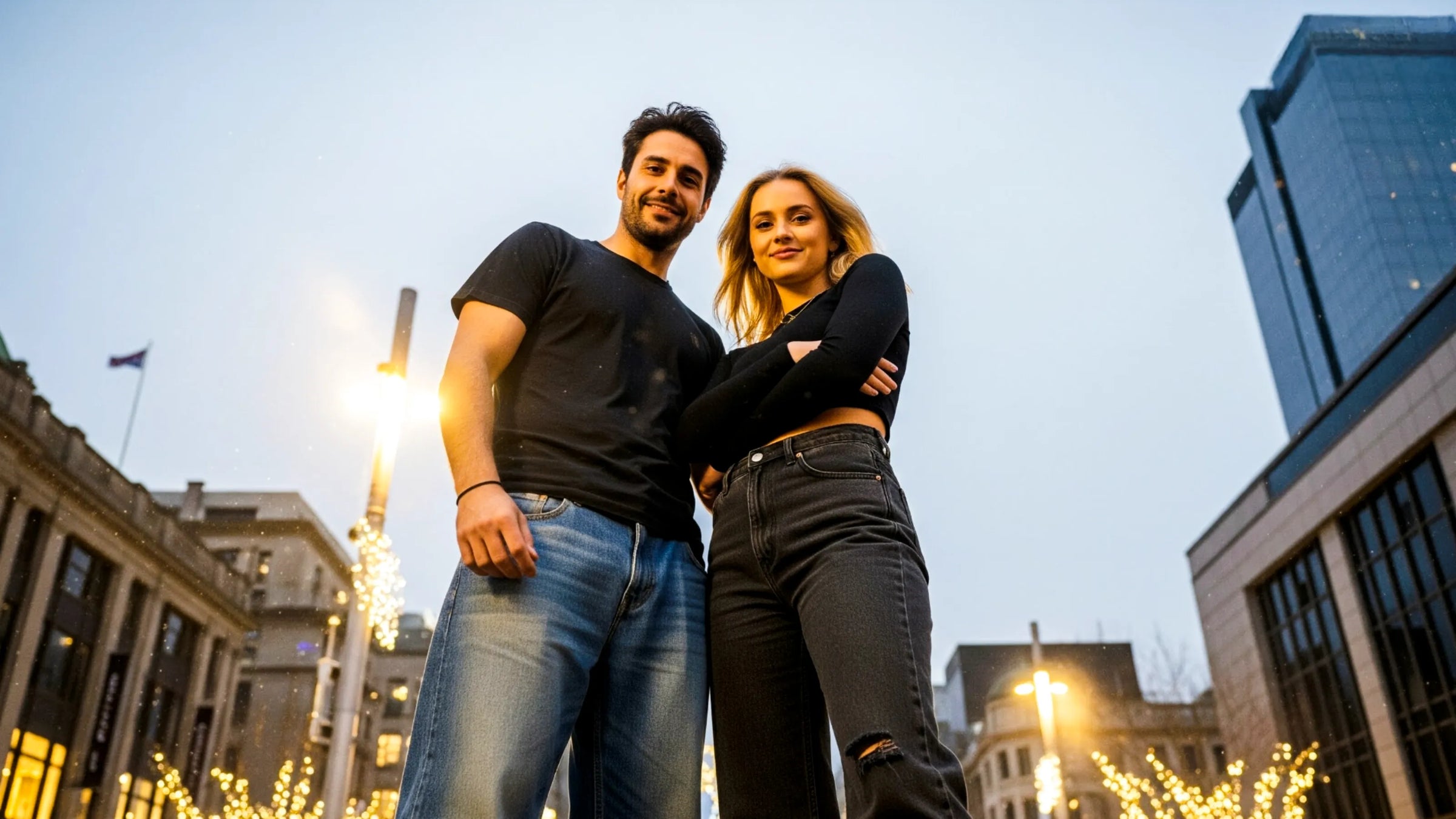 The image shows a man and woman standing confidently in an urban setting during the evening. The photo is taken from a low angle, making them appear tall and dominant against the sky. Both are wearing casual outfits—black tops and jeans. The background features modern buildings and decorative streetlights adorned with glowing string lights, giving the scene a festive yet stylish atmosphere, possibly around the holiday season or during a city event.
